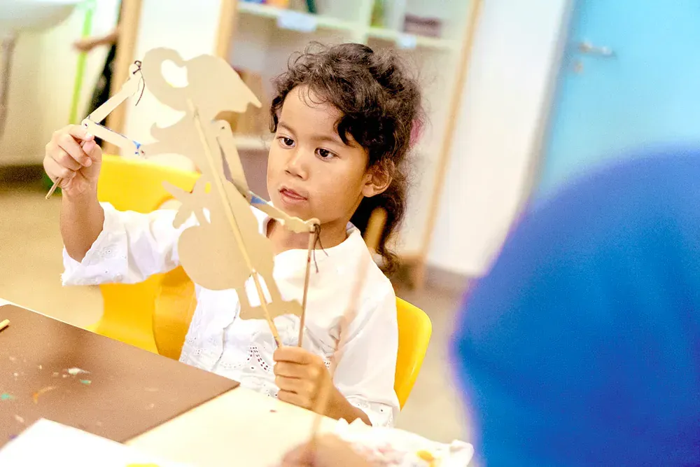 A little girl is sitting at a table holding a cardboard puppet.