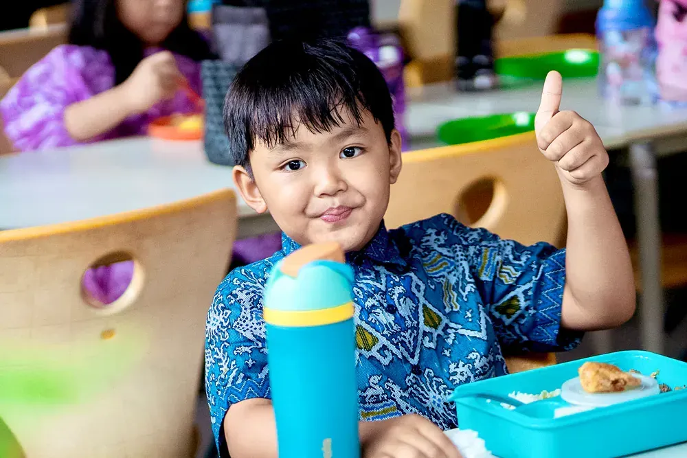 A young boy is sitting at a table with a lunch box and giving a thumbs up.