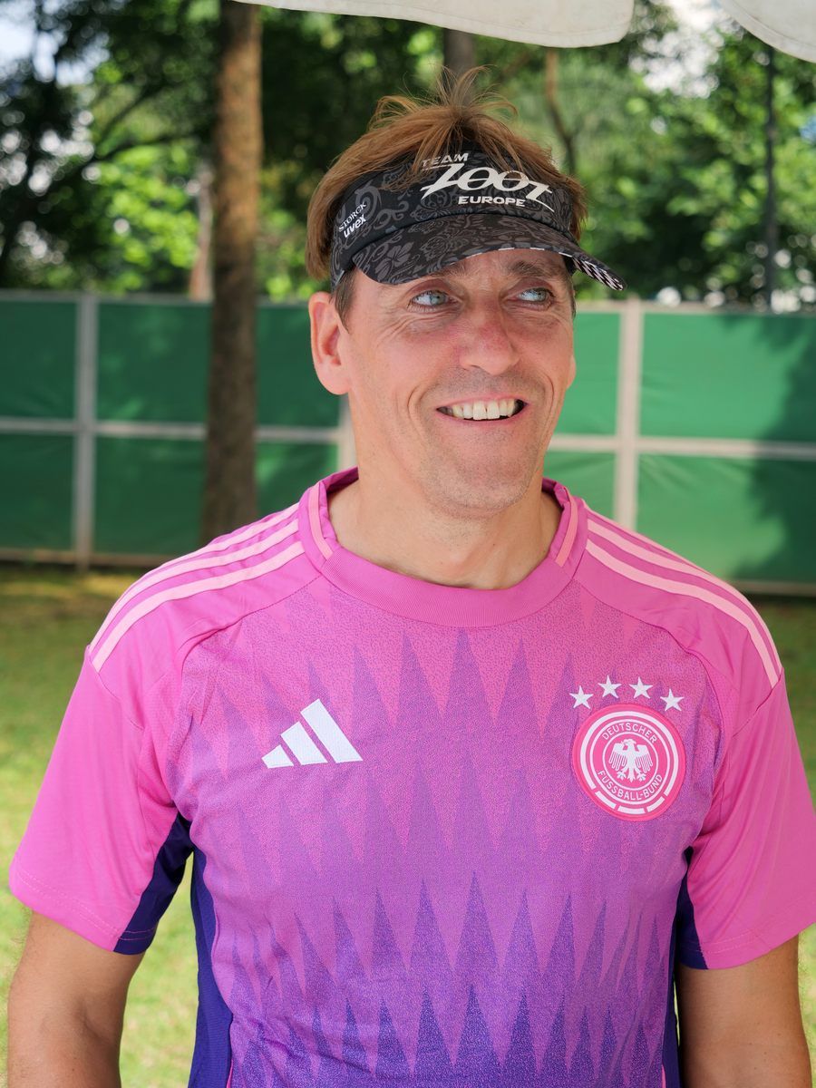 A man wearing a pink adidas shirt and a bandana on his head is smiling.