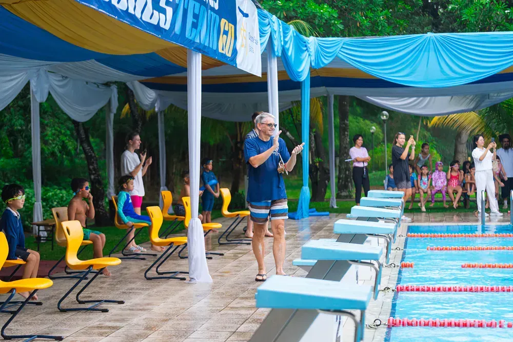 A man is standing in front of a swimming pool talking into a microphone.