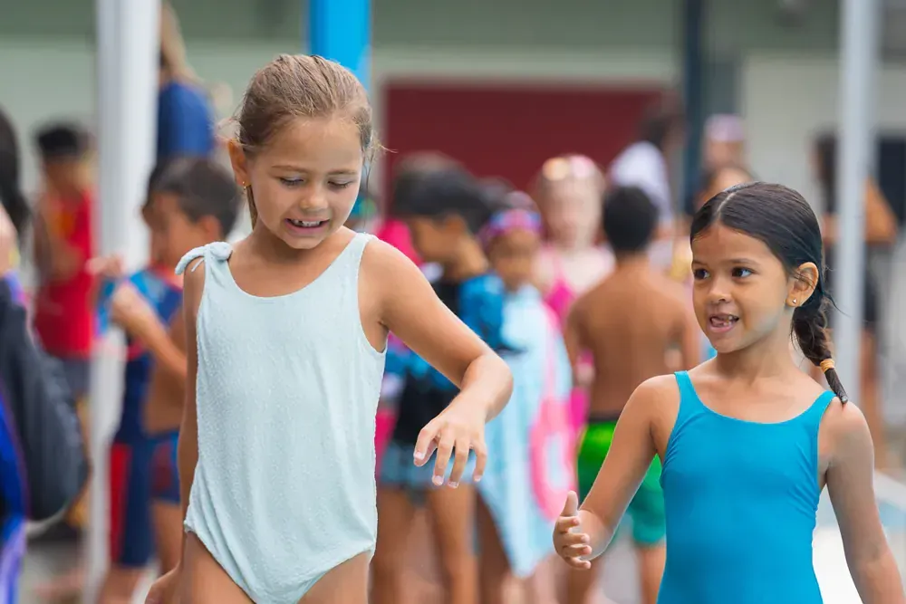 Two young girls in swimsuits are holding hands in front of a crowd of children.
