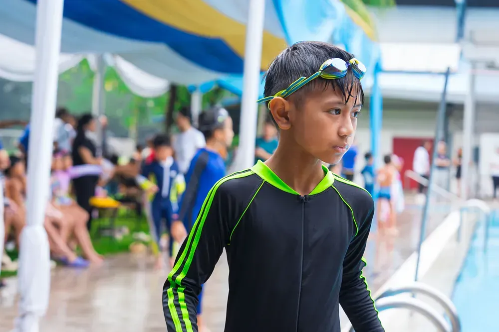 A young boy in a wet suit and goggles is standing next to a swimming pool.