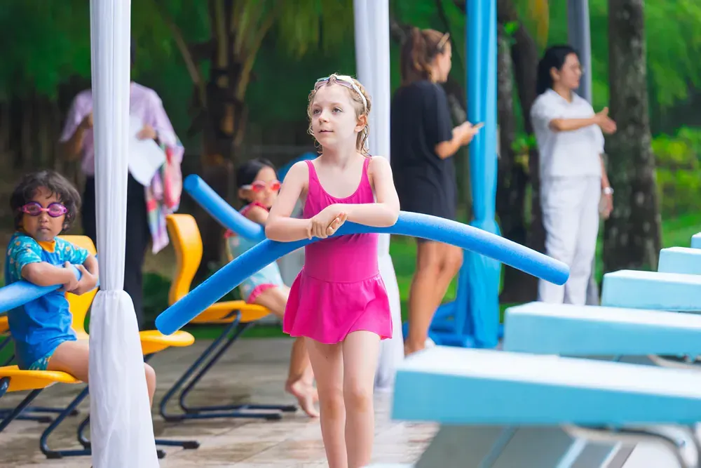 A little girl in a pink dress is holding a blue pool noodle.