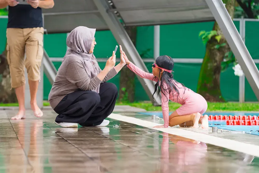 A woman and a little girl are giving each other a high five in front of a swimming pool.