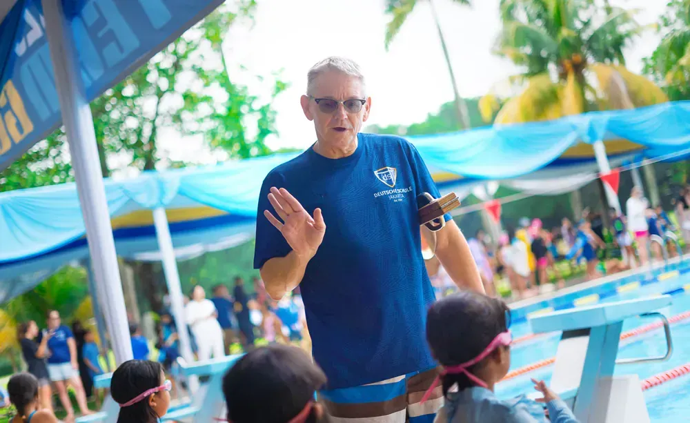 A man is standing in front of a swimming pool talking to children.