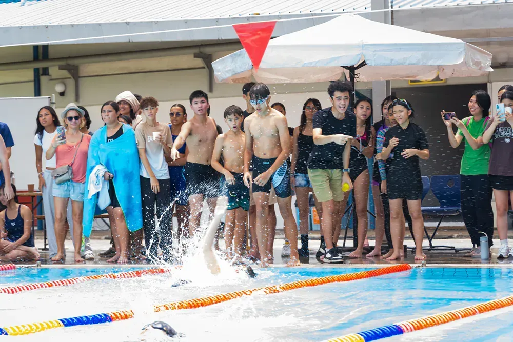 A group of people are standing around a swimming pool watching a boy swim.