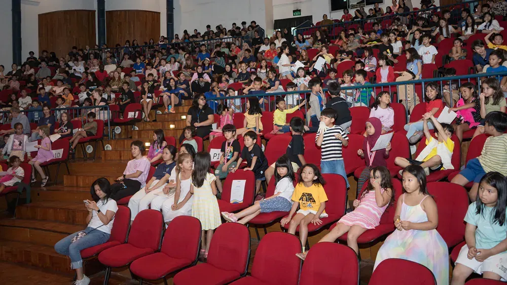 A large group of children are sitting in red chairs in an auditorium.