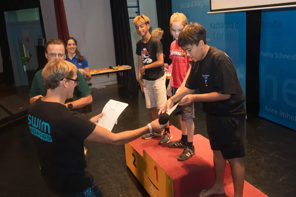 A man in a swim shirt is giving a certificate to a boy on a podium.