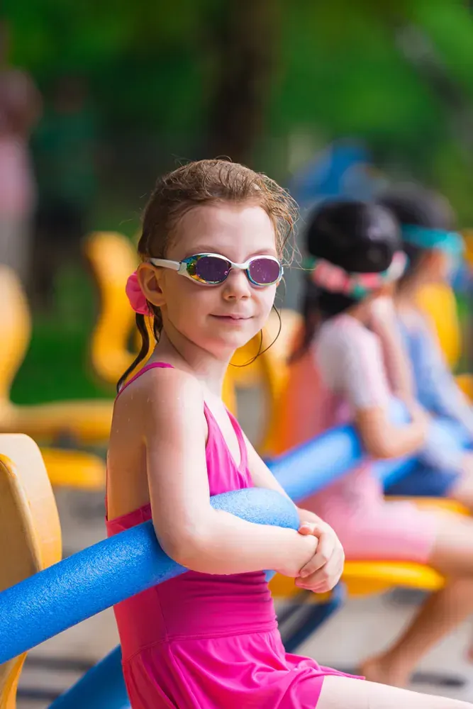 A little girl in a pink swimsuit and sunglasses is sitting on a bench holding a pool noodle.