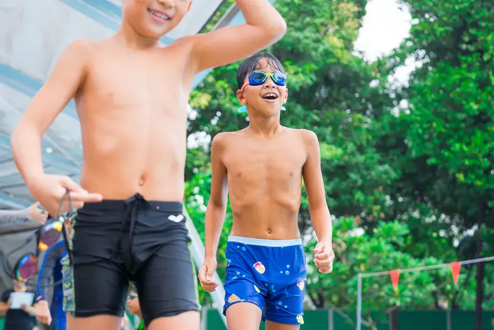 Two boys in swimming trunks. The boy in blue swimming trunks is wearing blue googles and he is smilling.