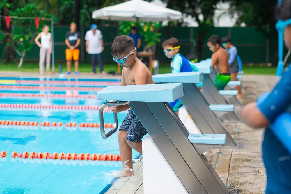 A group of young boys are sitting on starting blocks at a swimming pool.