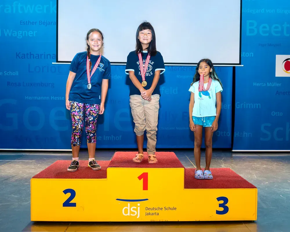 Three young girls are standing on a yellow podium with the number 1 on it