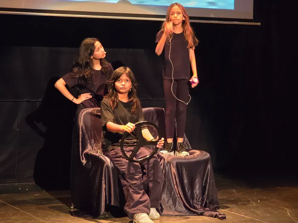 Three girls are standing on a stage and one is holding a steering wheel