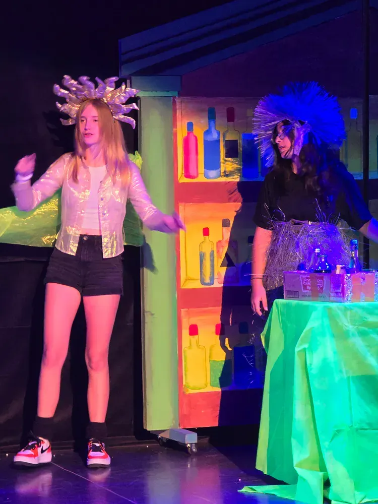Two women are standing on a stage in front of a shelf with bottles on it.