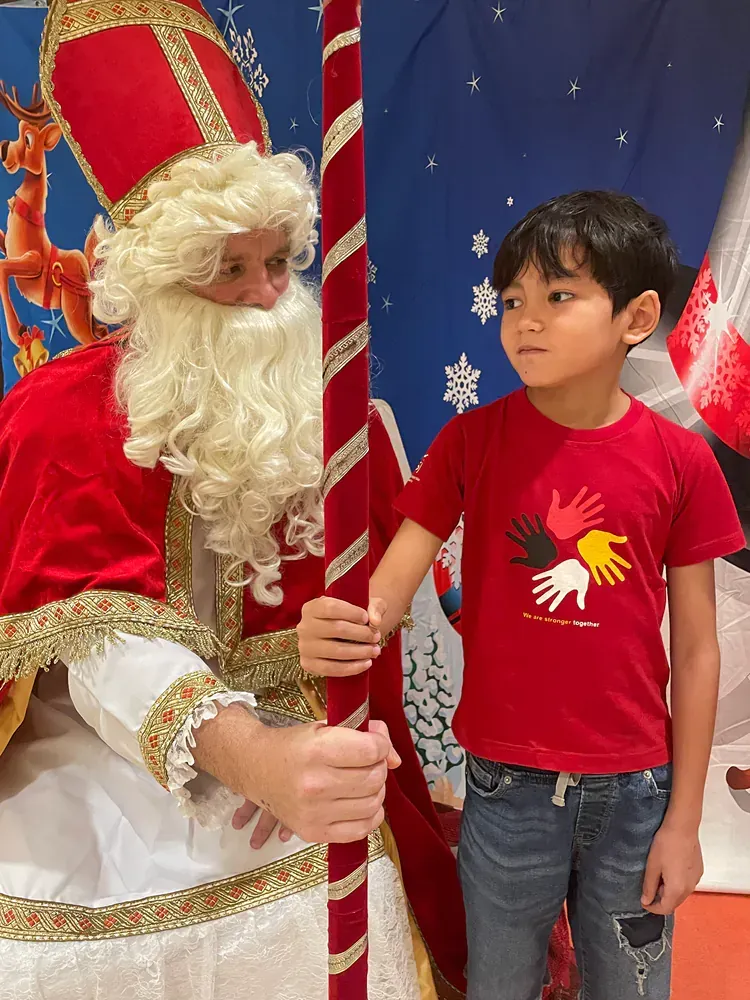 A boy in a red shirt is standing next to a man dressed as santa claus.