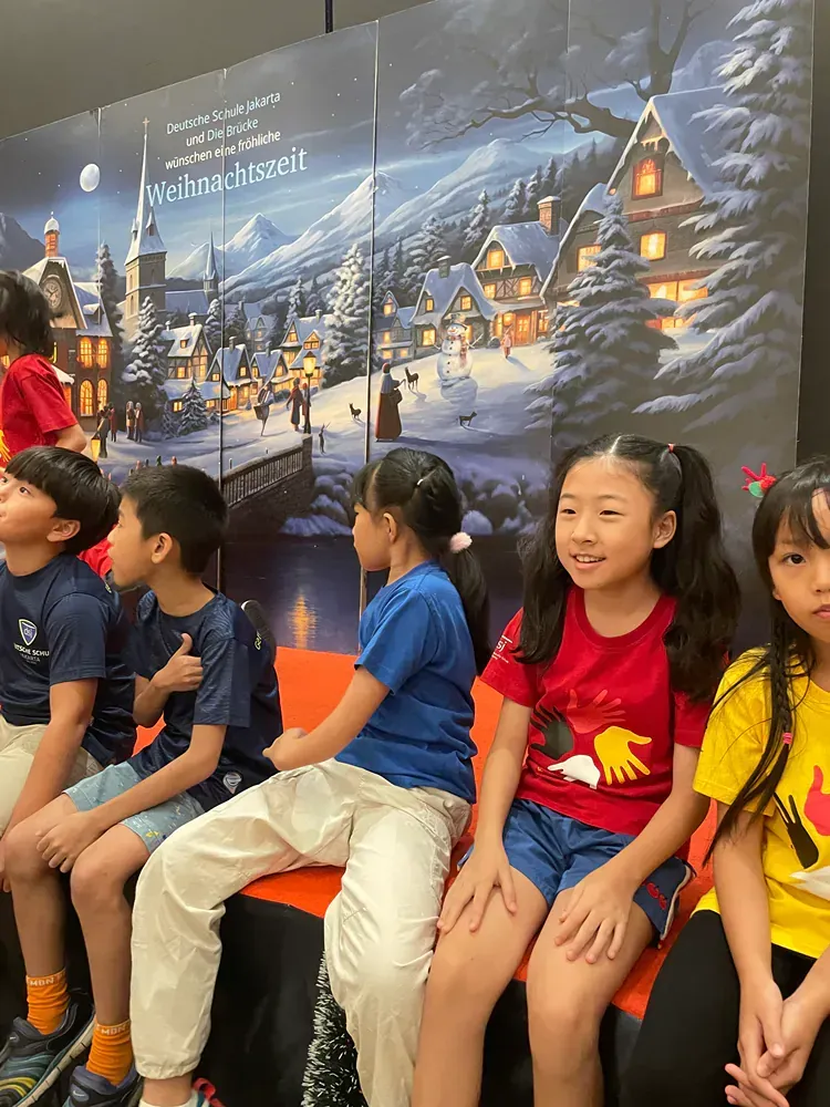 A group of children are sitting in front of a painting of a snowy village.