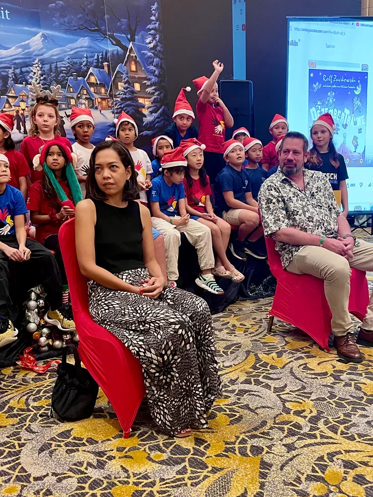 A group of people are sitting in red chairs in front of a group of children wearing santa hats.