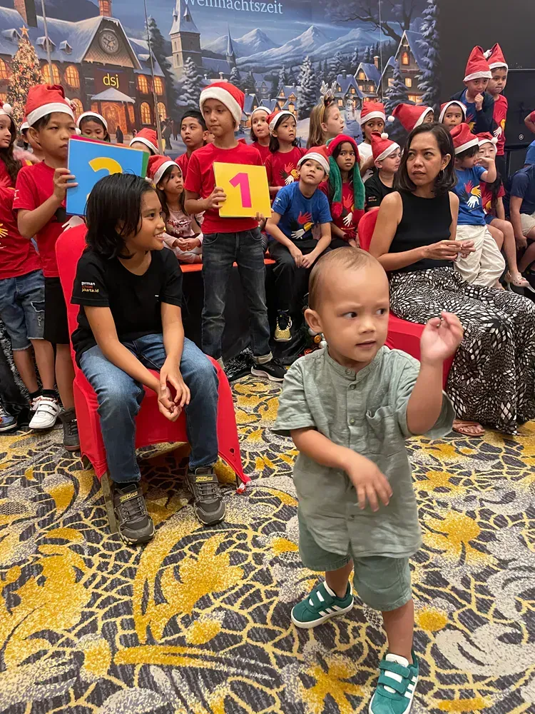 A baby is standing in front of a group of children wearing santa hats.