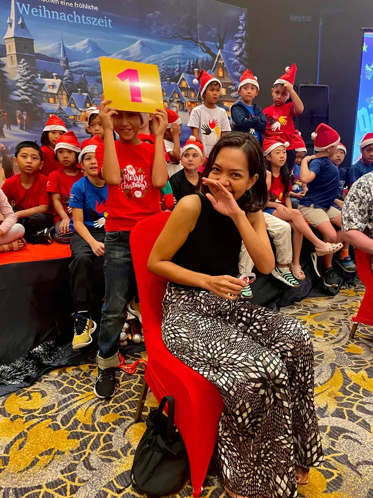 A woman is sitting in a red chair in front of a group of children wearing santa hats.