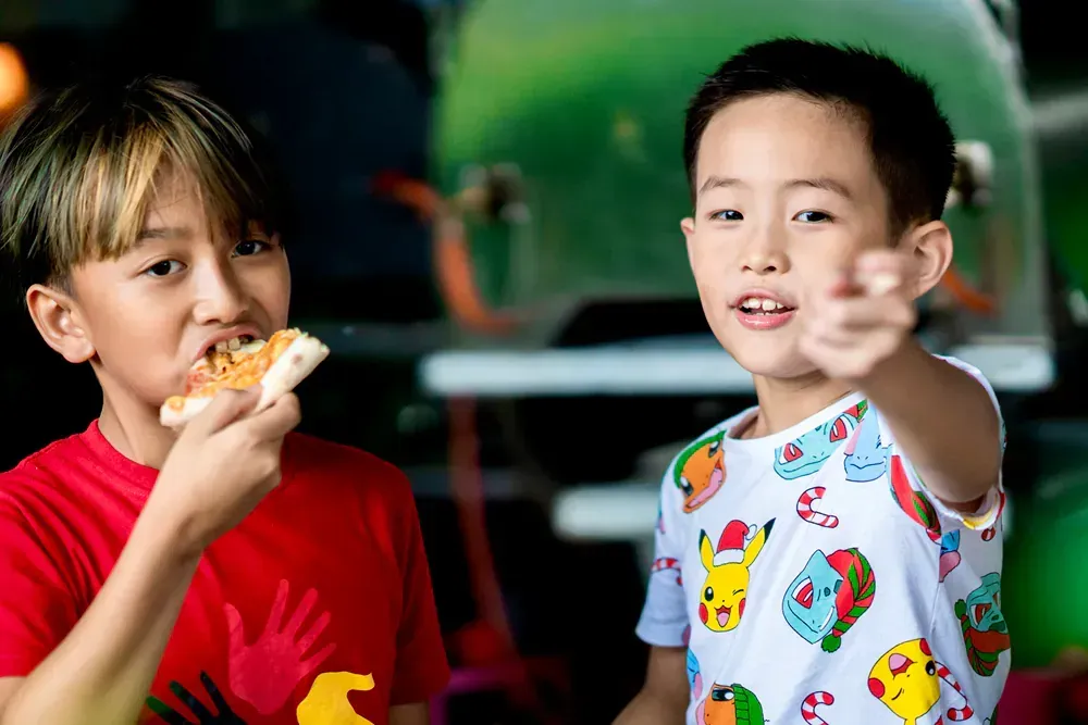 Two young boys are eating pizza and one of them is pointing at the camera.