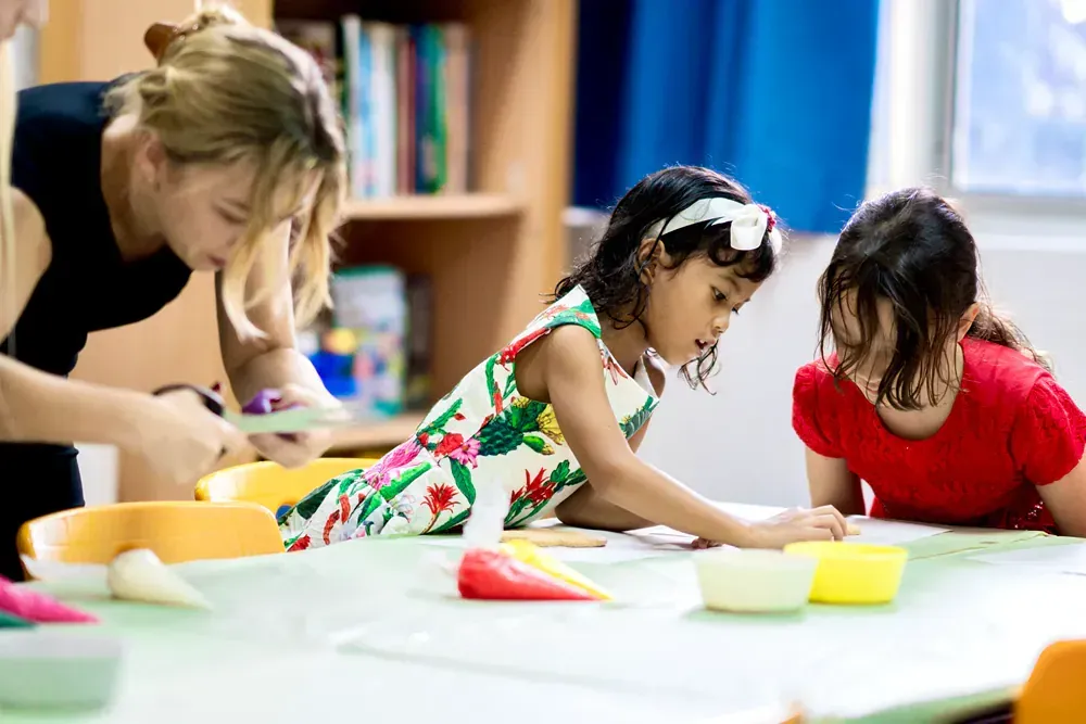 A woman is teaching two little girls how to use scissors at a table.