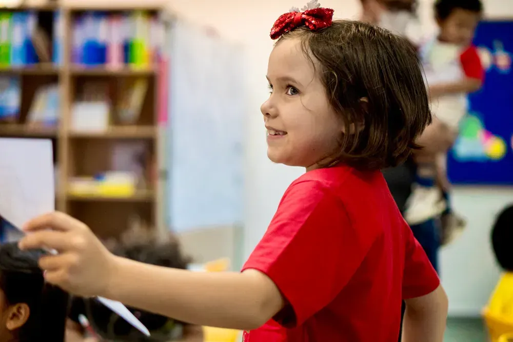 A little girl in a red shirt is standing in a classroom.