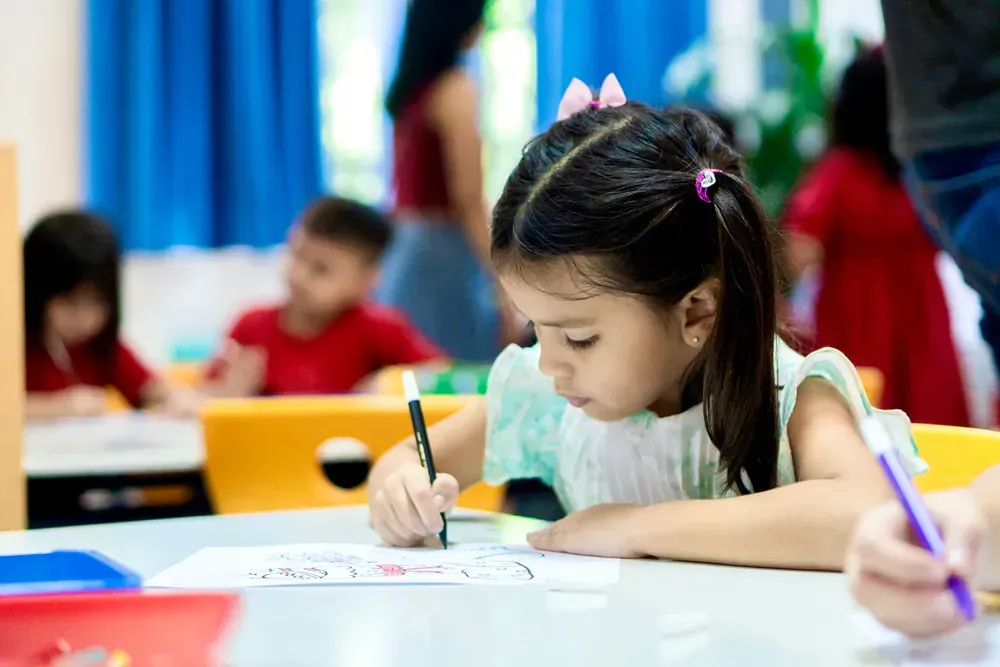 A little girl is sitting at a table in a classroom writing on a piece of paper.