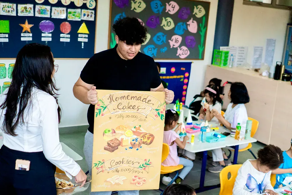 A man is holding a sign in front of a group of children in a classroom.