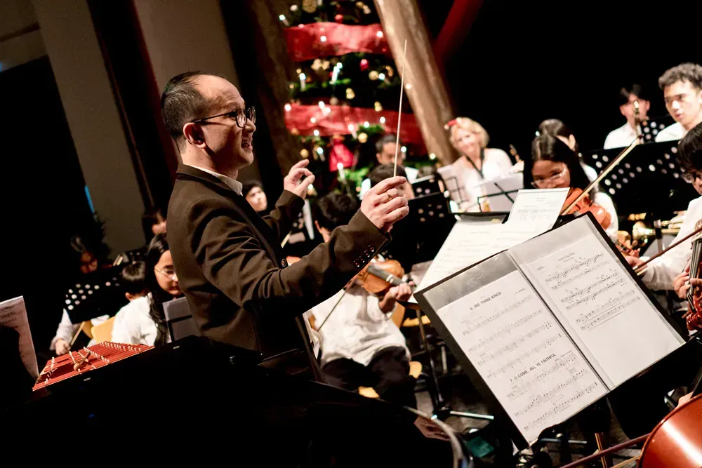 A man is leading an orchestra on a stage with a christmas tree in the background.