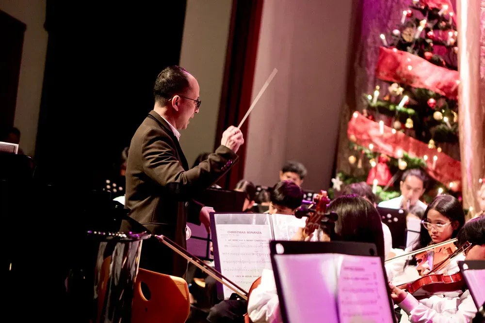 A man is leading an orchestra on a stage with a christmas tree in the background.