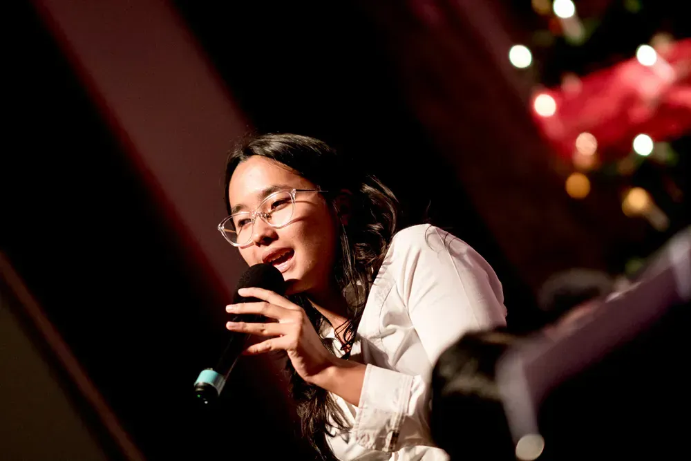 A woman is singing into a microphone on a stage in front of a christmas tree.