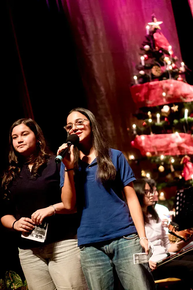 Two girls are singing into microphones on a stage in front of a christmas tree.
