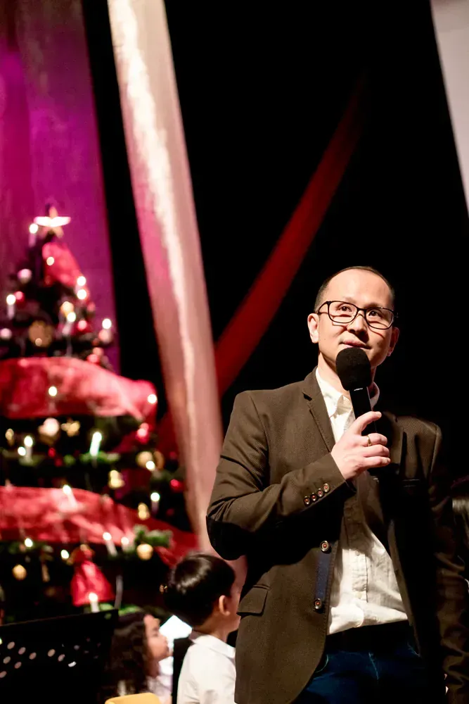 A man is standing on a stage holding a microphone in front of a christmas tree.