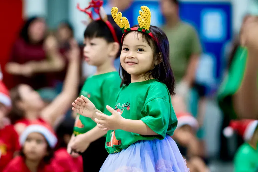 A little girl wearing a reindeer headband is standing in front of a crowd of children.