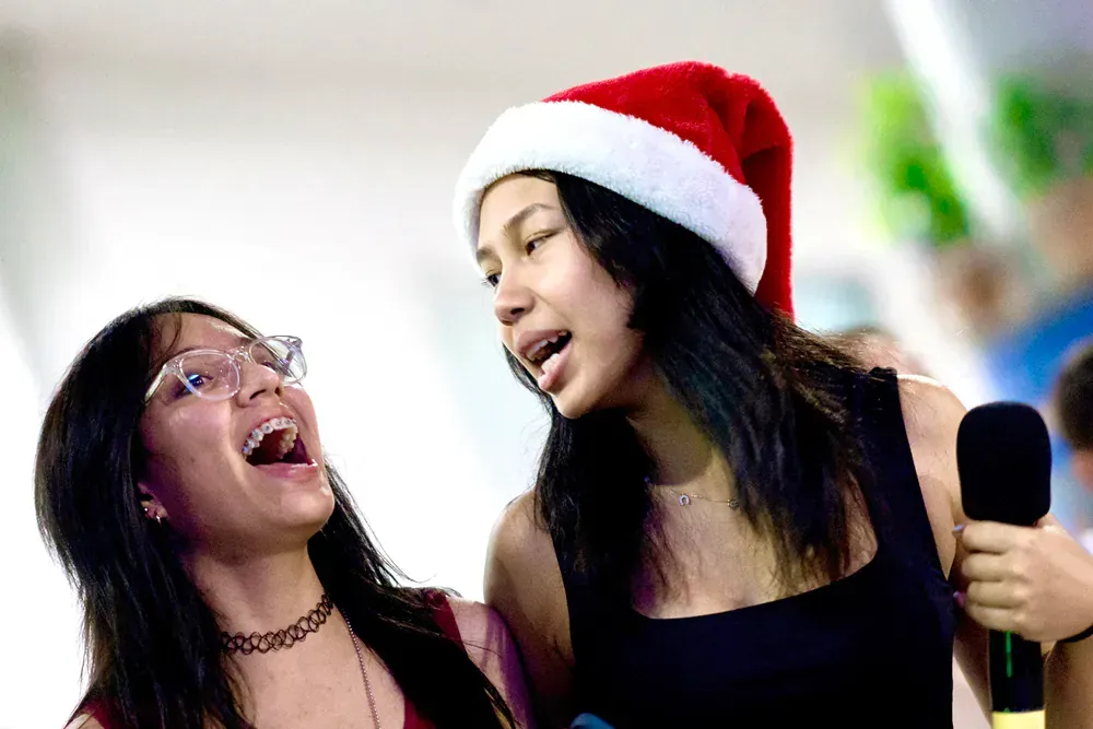Two girls wearing santa hats are singing into microphones.