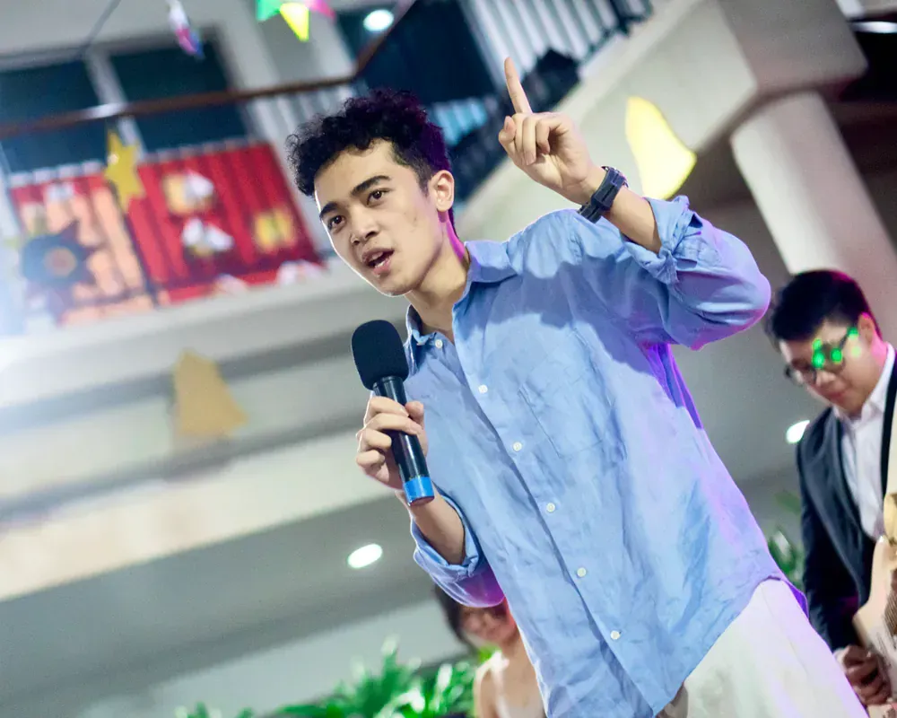 A man in a blue shirt is holding a microphone and pointing up.