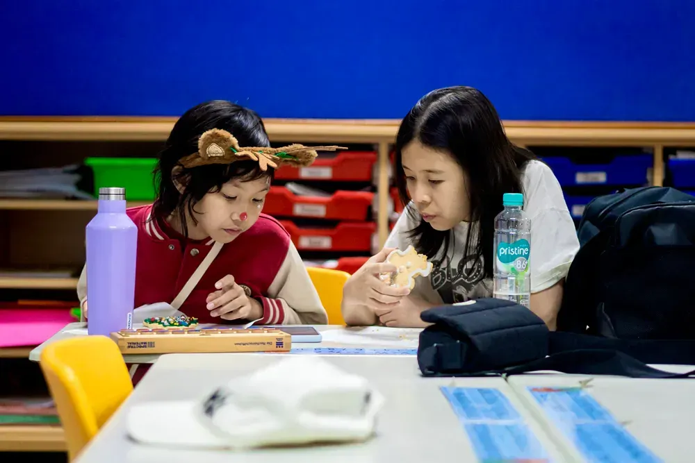 Two young girls are sitting at a table in a classroom eating sandwiches.