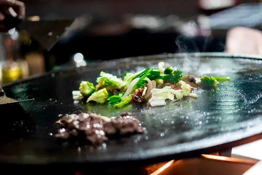A person is cooking meat and vegetables on a pan.