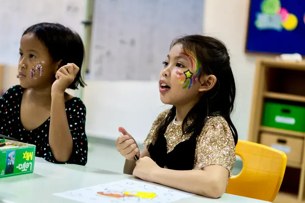 Two little girls are sitting at a table with their faces painted.