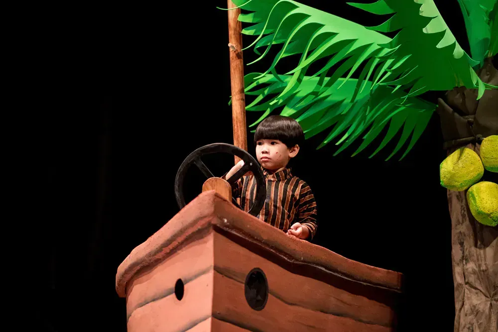 A little boy is sitting in a wooden boat with a steering wheel.