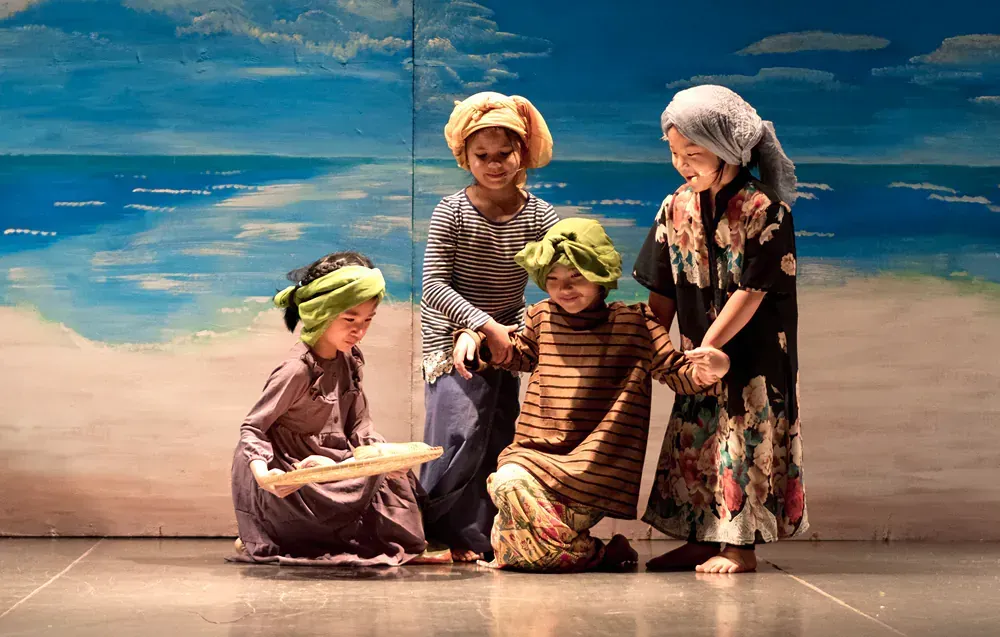 A group of children are standing on a stage in front of a painting of the ocean.