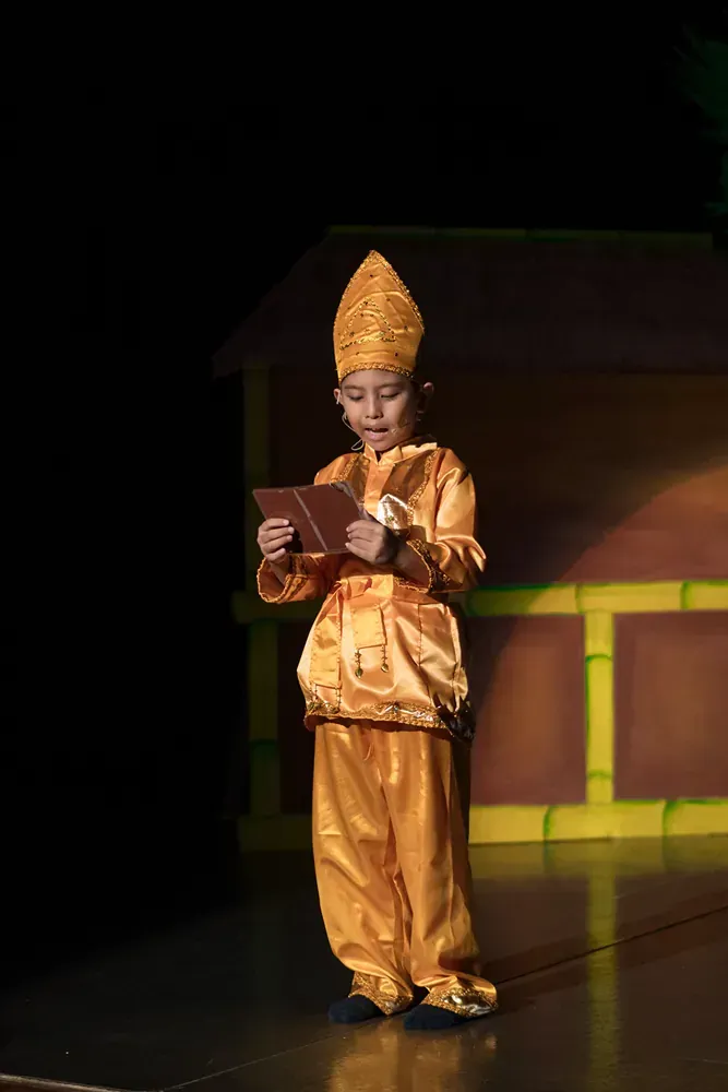 A young boy in a gold costume is standing on a stage reading a book.