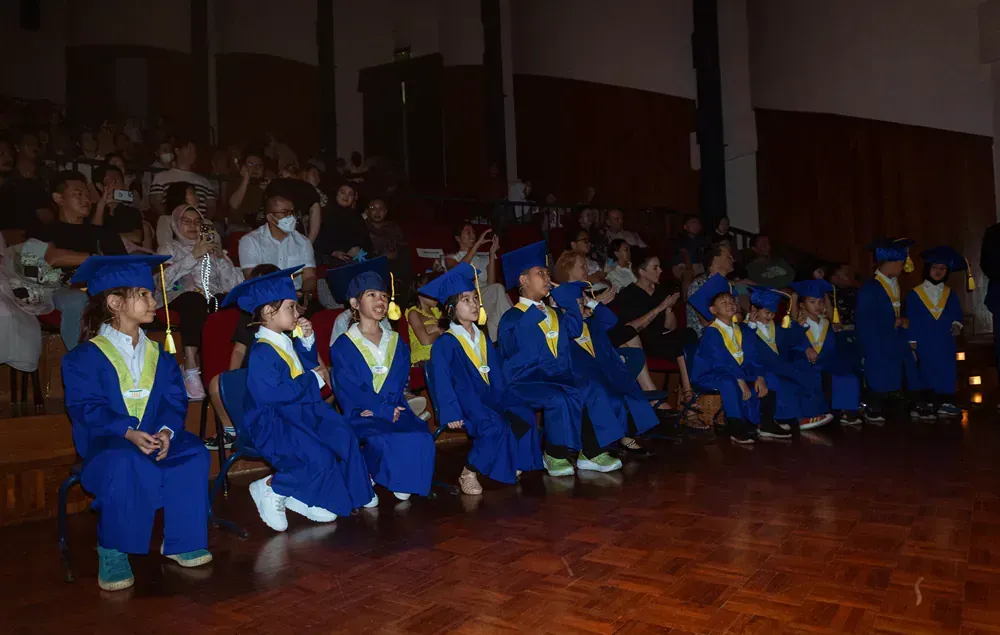 A group of children in graduation gowns and caps are sitting on the floor in front of a crowd.