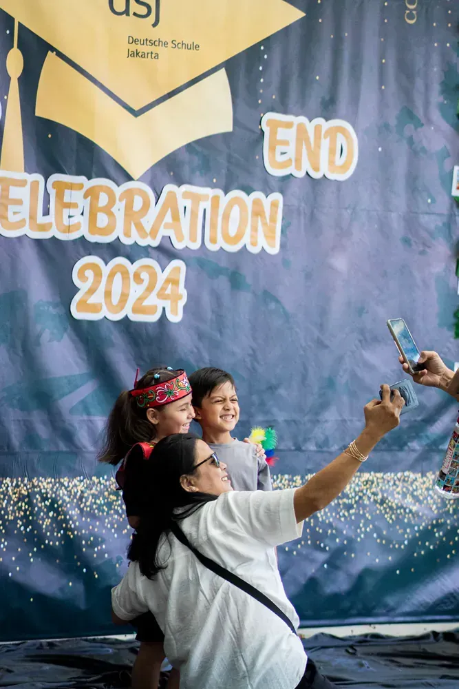 A woman is taking a selfie with two children in front of a graduation banner.