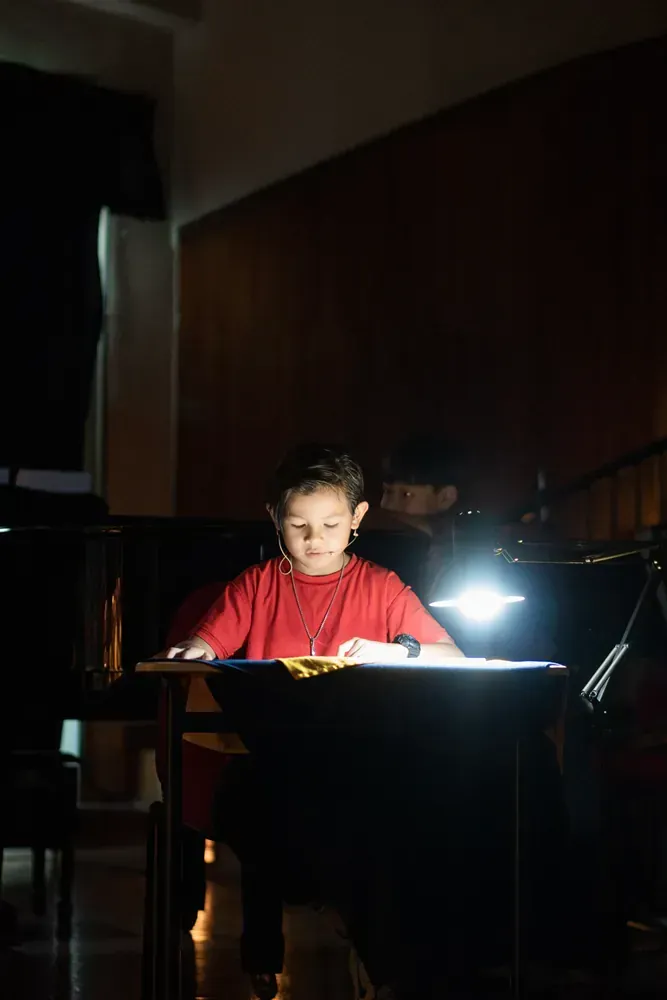 A young boy is sitting at a desk in the dark reading a book.