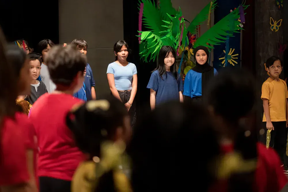 A group of children are standing on a stage in front of a green bird.