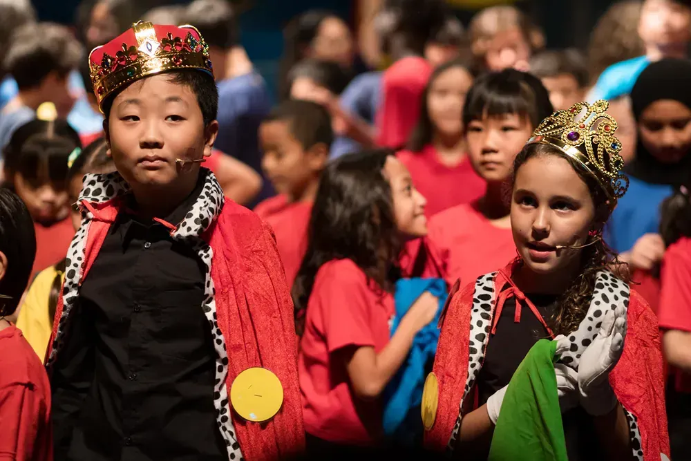 A boy and a girl wearing crowns are standing in front of a crowd of children.
