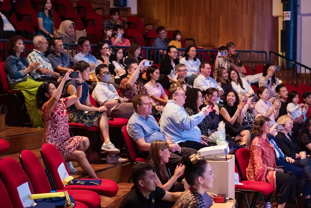 A large group of people are sitting in red chairs in an auditorium.