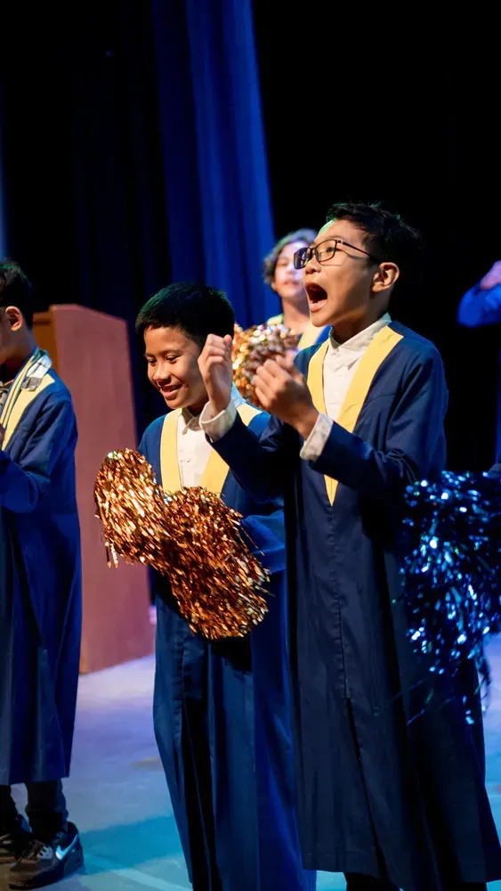 A group of young boys in graduation gowns are standing on a stage holding pom poms.