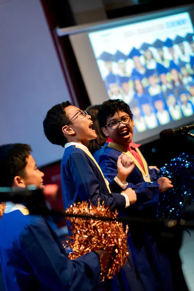 A group of boys in graduation gowns are singing in front of a projector screen.
